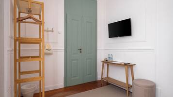 Une pièce avec une échelle en bois, une porte verte, un téléviseur fixé au mur, une petite table en bois avec des bouteilles et un pouf rond.