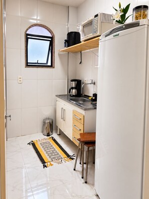 Eine kleine Küche mit Mikrowelle, Kaffeemaschine und einem Kühlschrank. Ein Fenster bietet einen Ausblick, ein Mülleimer steht bereit und ein gemustertes Teppich liegt auf dem Boden.
