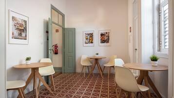 Une pièce avec un sol à motifs, des murs blancs et une porte qui donne sur une autre pièce. On y trouve deux tables rondes avec des chaises, ainsi que deux tableaux encadrés accrochés au mur.