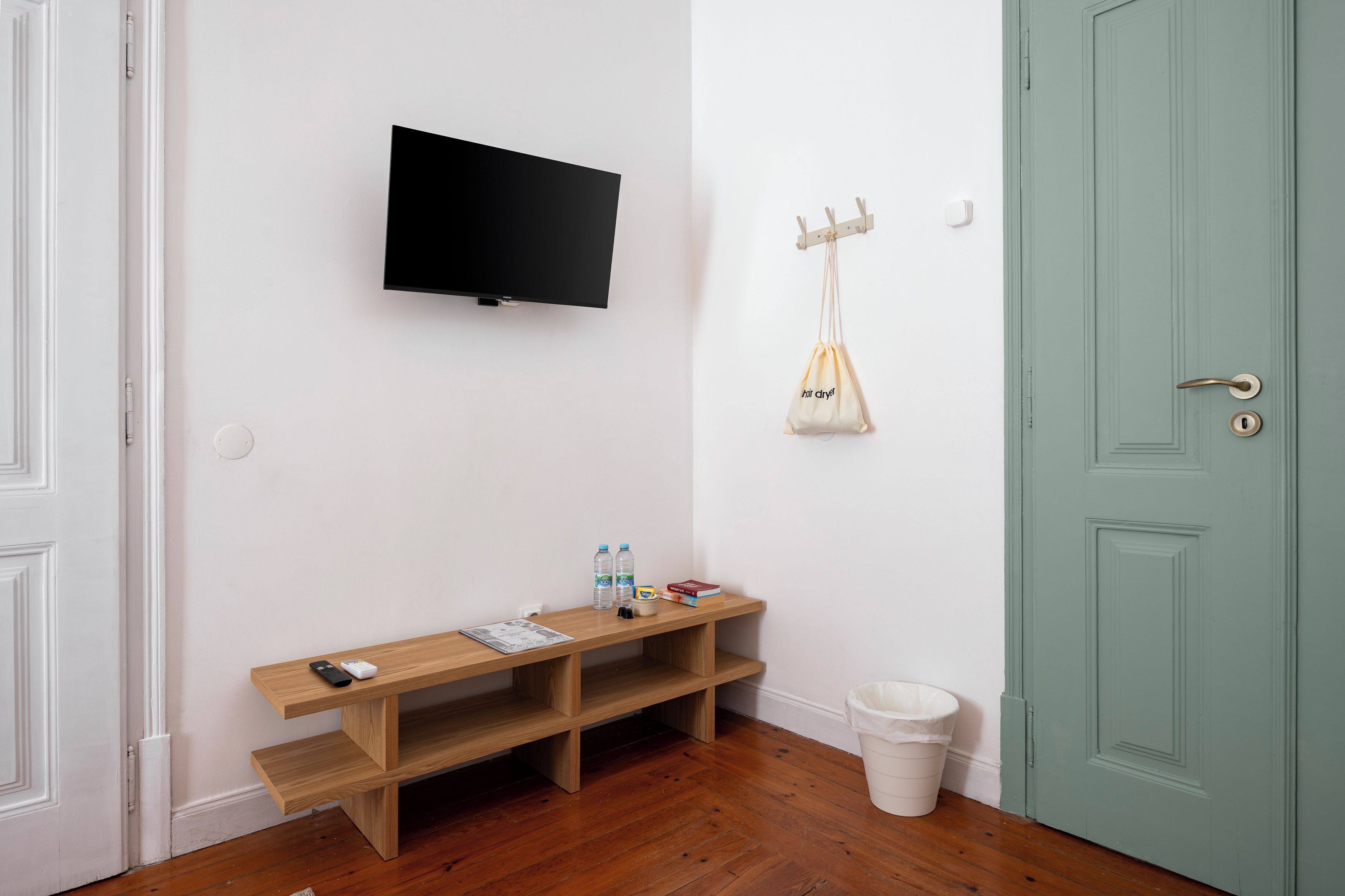 Una stanza con una TV a schermo piatto montata a parete, una panca di legno con una rivista, due bottiglie d'acqua, un libro e un cestino bianco.