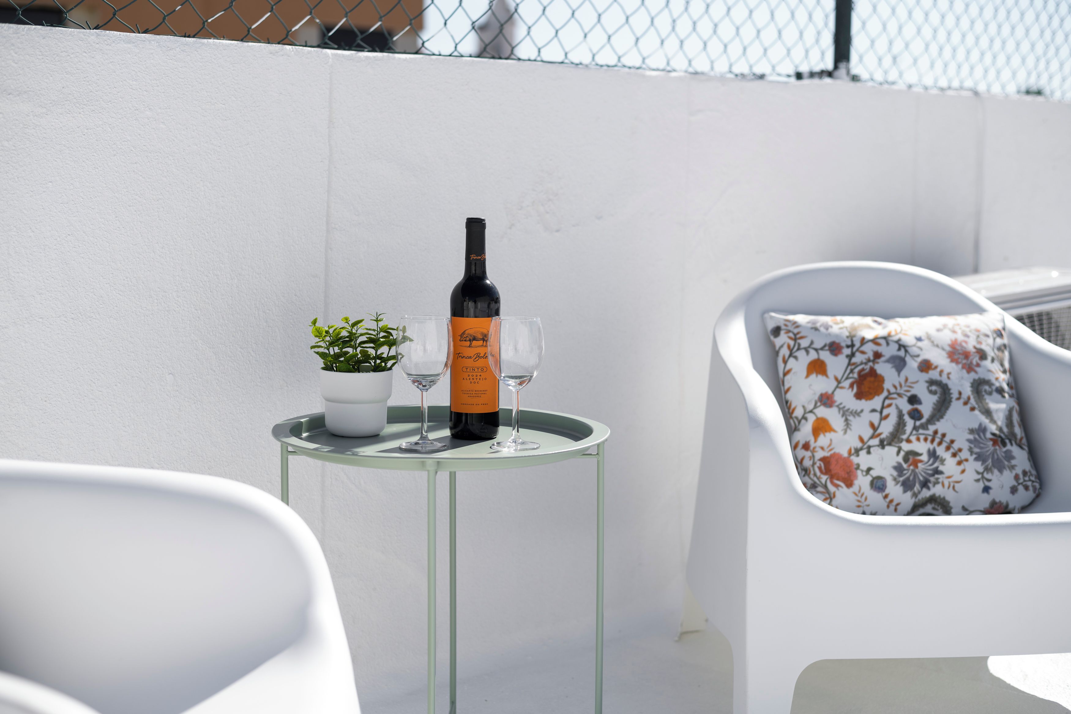 Une table en verre avec une bouteille et deux verres à vin, une plante en pot et une chaise blanche agrémentée d’un coussin à motifs floraux.