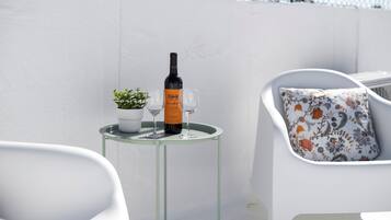Une table en verre avec une bouteille et deux verres à vin, une plante en pot et une chaise blanche agrémentée d’un coussin à motifs floraux.