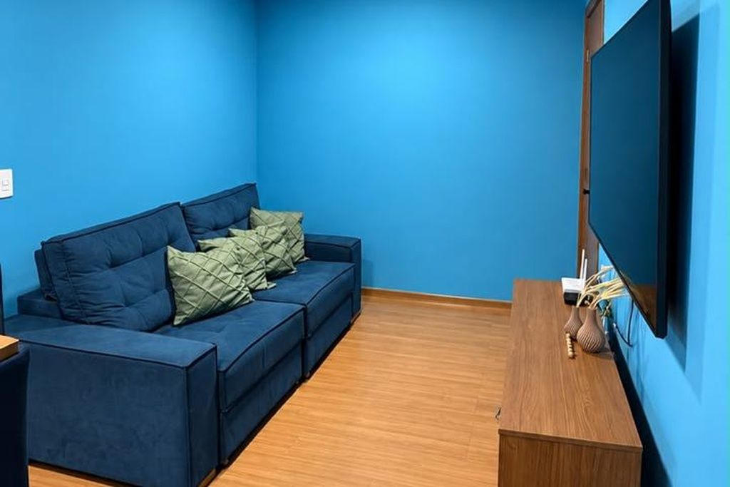 A room with a blue wall, a wooden floor, a blue sofa with cushions, and a flat-screen TV mounted on the wall.