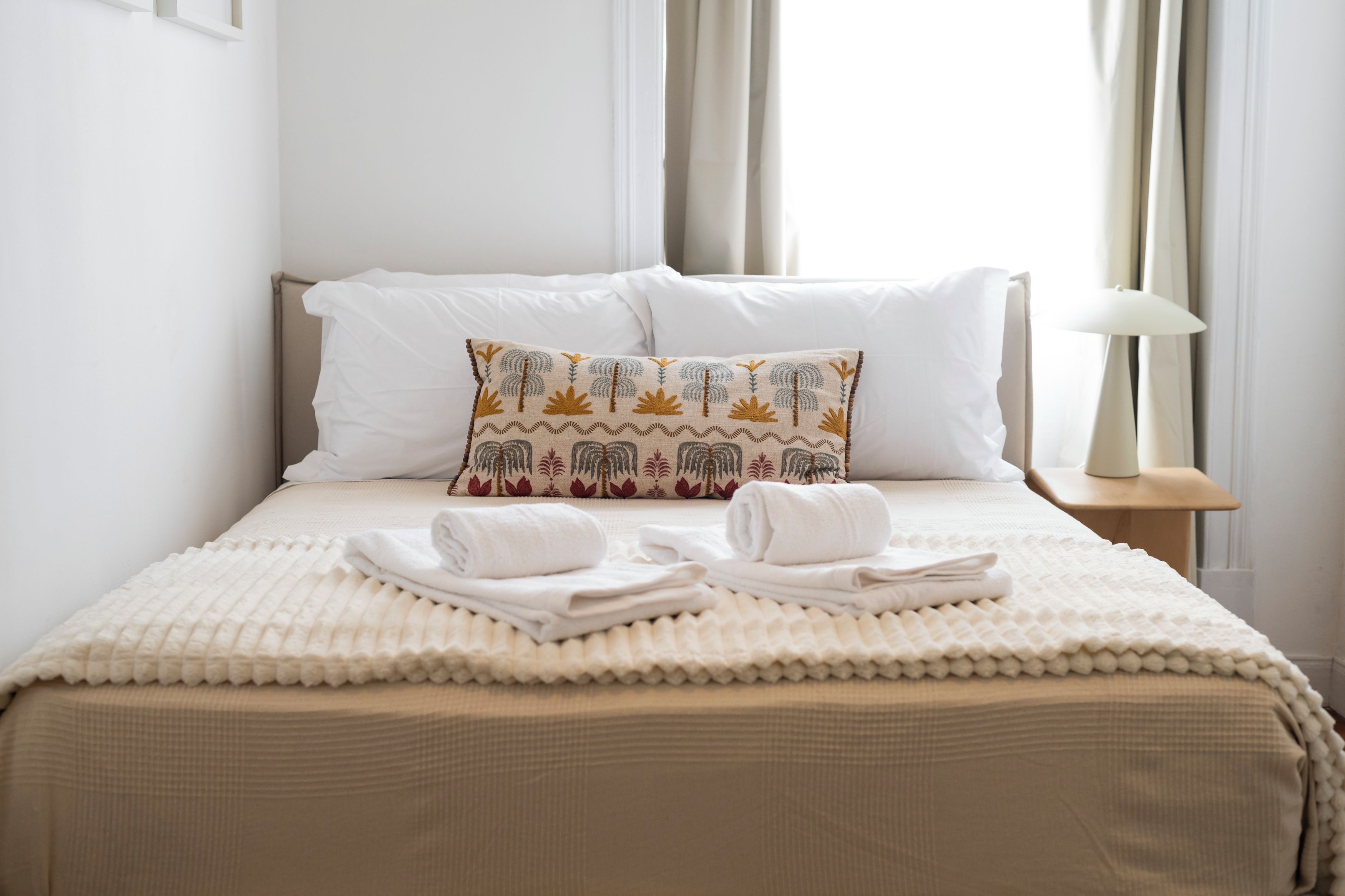 A neatly made bed with a patterned pillow, a folded towel, and a bedside lamp on a wooden nightstand.