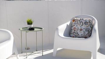 Una silla blanca con un cojín floral, una mesita redonda con una planta en maceta y una pared blanca.