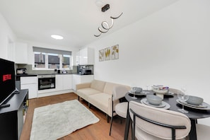 A modern kitchen with white cabinets, a dining area with a beige sofa, and a flat-screen TV on the left. - Hamdan Suites Stratford (London)