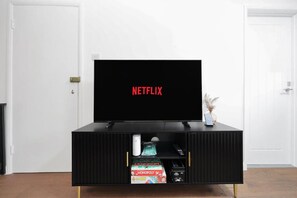 A black entertainment unit with a flat-screen TV displaying 'NETFLIX' in red letters, a remote control, and a small vase with dried plants. - Hamdan Suites Stratford (London)