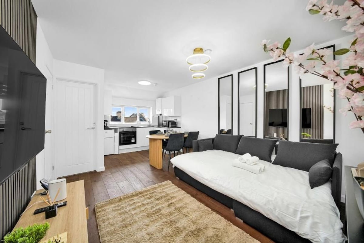 A modern bedroom with a large bed, a dining area, and a kitchen in the background.