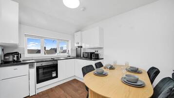Una cocina moderna con armarios blancos, horno empotrado, una mesa de comedor con sillas y una ventana con vistas a edificios.