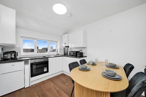 A modern kitchen with white cabinetry, a built-in oven, a dining table with chairs, and a window with a view of buildings. - Hamdan Suites Stratford (London)