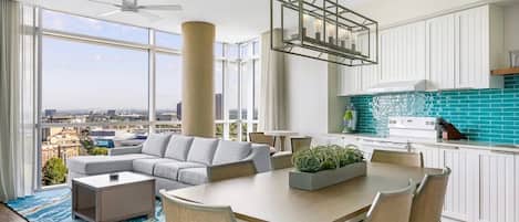 Un salon moderne avec un canapé, une table basse et une table à manger, une cuisine avec des meubles blancs et un pare-feu turquoise, et une grande fenêtre offrant une vue sur la ville.