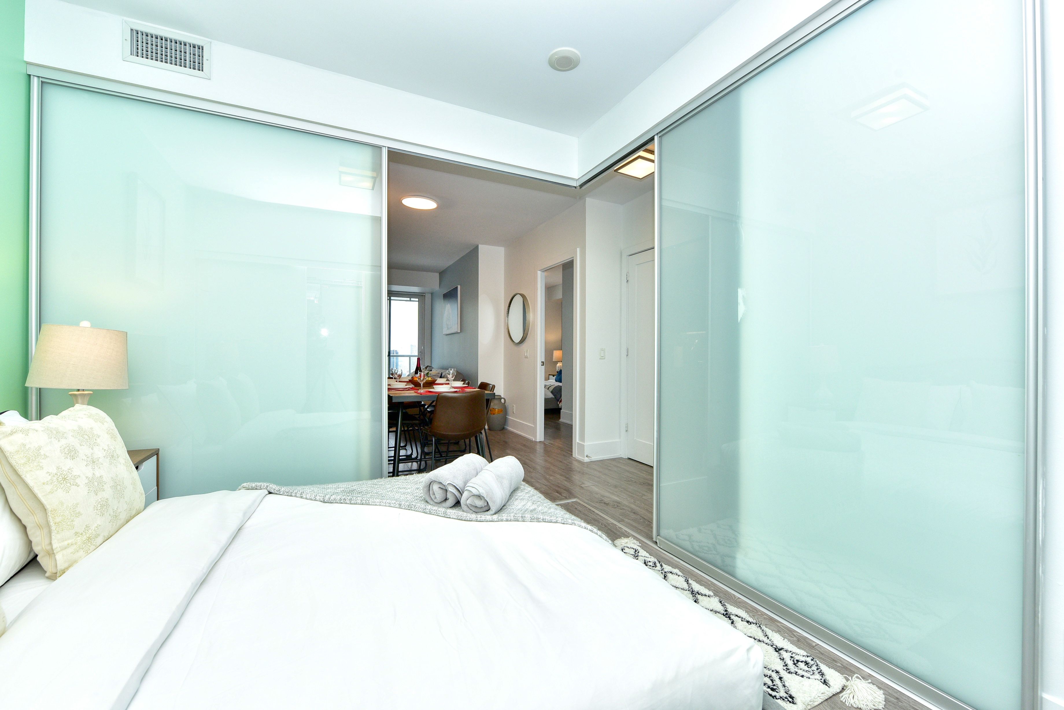 Quarto de hotel moderno com uma cama grande, uma mesa de escritório e uma área de refeições visível através de um painel de vidro.