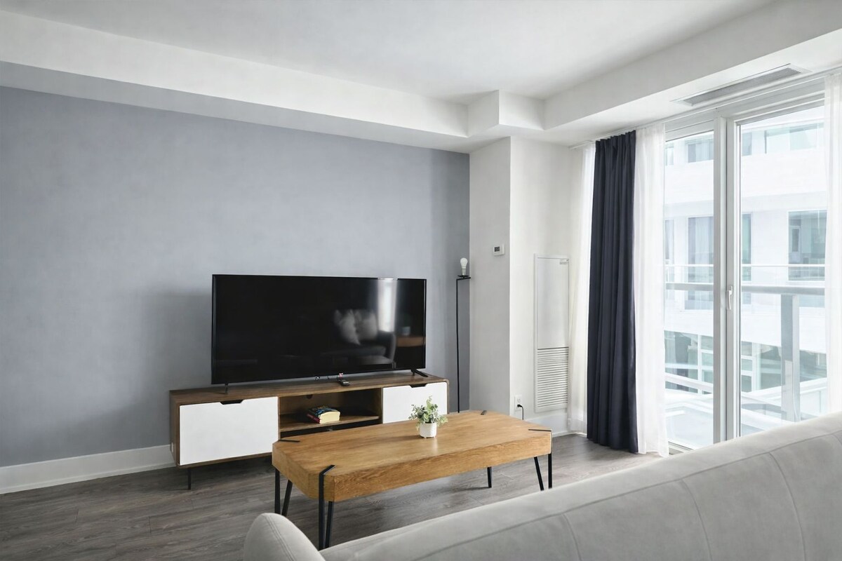 Un salon moderne avec un téléviseur à écran plat, une table basse en bois, un canapé et une grande fenêtre avec des rideaux.