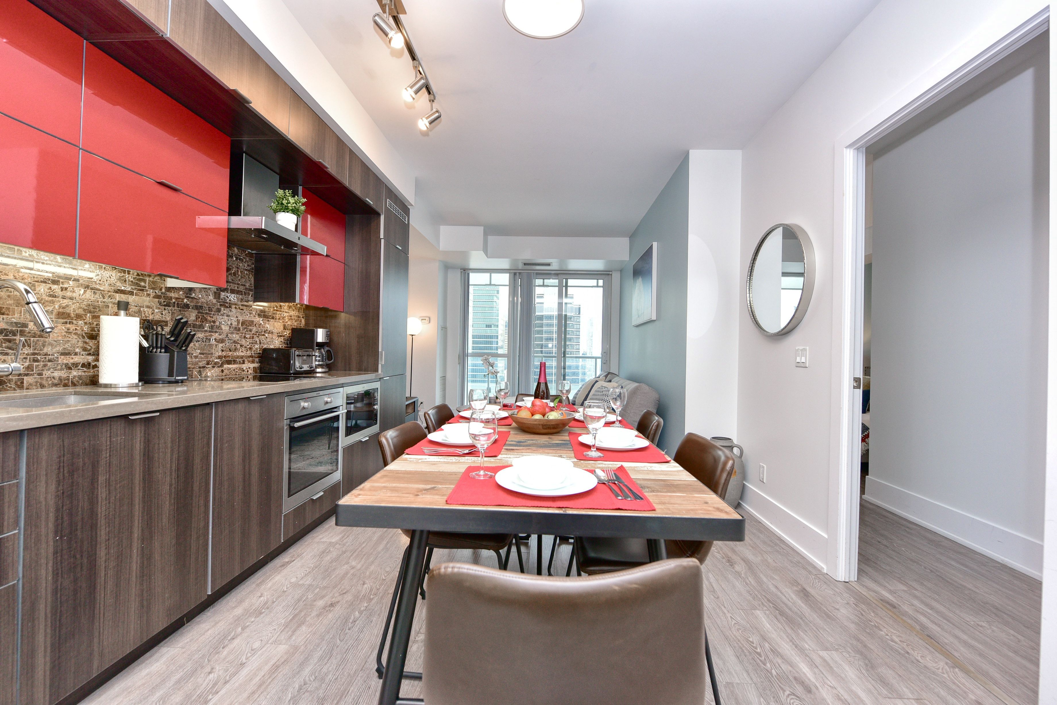 Une cuisine moderne avec des meubles rouges, un coin repas doté d’une table en bois pour quatre personnes, et une vue sur la ville grâce à de grandes fenêtres.