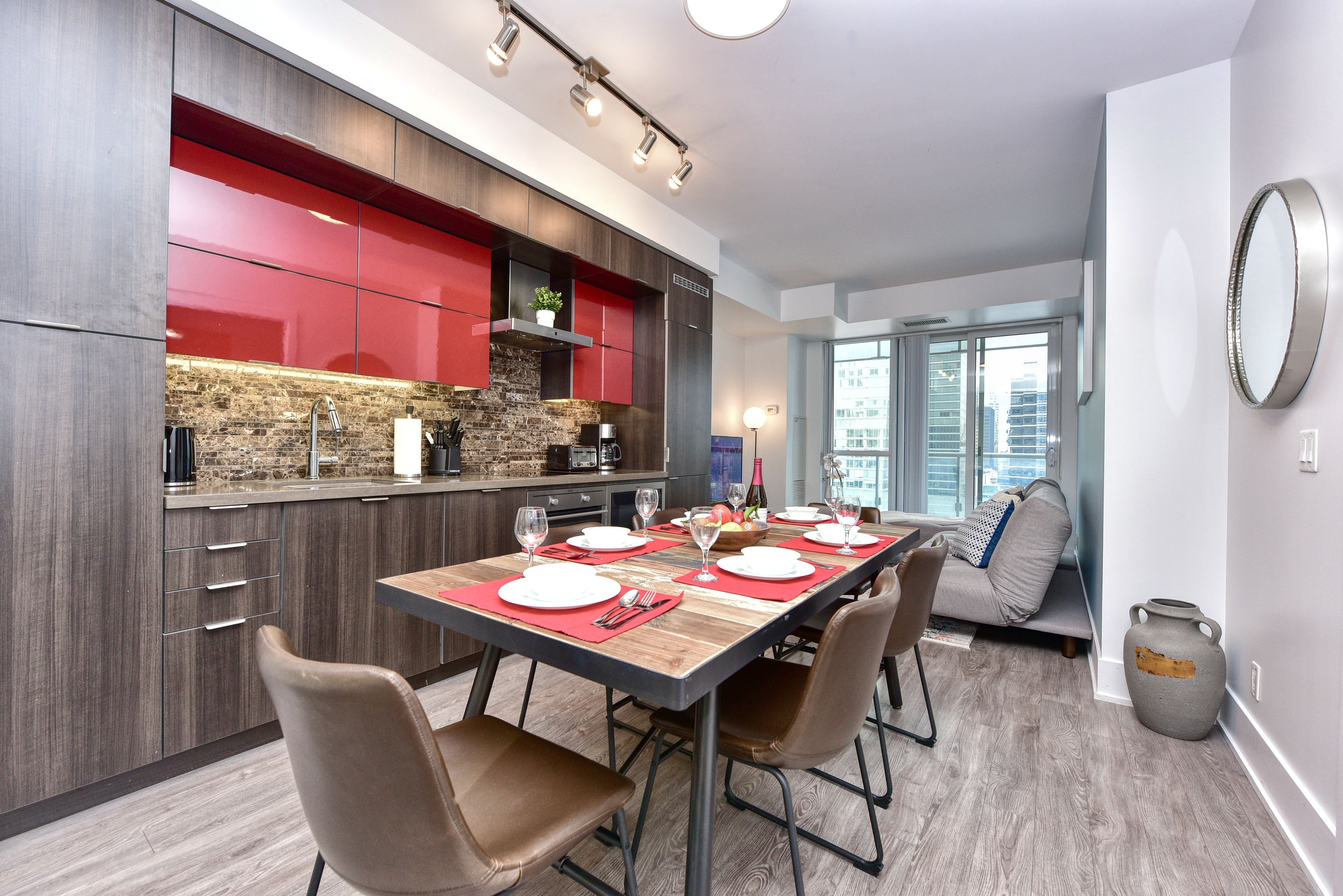 A modern kitchen with red cabinets, a wooden dining table set for four, and a large window offering a view of the city.