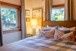 A bedroom with a bed, curtains, a lamp, and framed pictures on the wall. - Cannon Beach Cottage – Walk to Beach & Downtown, Fenced Yard + Dog Friendly (Cannon Beach)