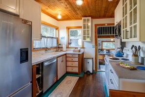 A kitchen with wooden cabinets, a stainless steel refrigerator, and a window overlooking a garden. - Cannon Beach Cottage – Walk to Beach & Downtown, Fenced Yard + Dog Friendly (Cannon Beach)