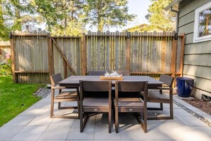 Eine Terrasse im Freien mit einem Holztisch und -stühlen, einem Holzzaun im Hintergrund und einem Haus zur Rechten.