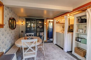 A compact kitchen with a dining area, featuring a round table, chairs, and a cabinet with glass doors. - Cannon Beach Cottage – Walk to Beach & Downtown, Fenced Yard + Dog Friendly (Cannon Beach)