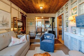 A cozy living room with a wooden ceiling, a sofa, a blue armchair, and a fireplace. - Cannon Beach Cottage – Walk to Beach & Downtown, Fenced Yard + Dog Friendly (Cannon Beach)
