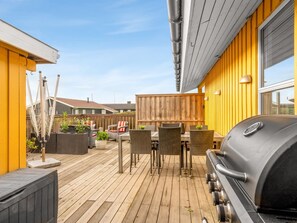 Een houten terras met buitenmeubilair, een barbecue en een gele muur.
