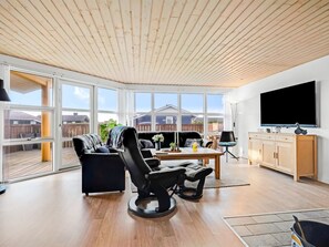 A modern living room with a wooden ceiling, large windows, a black leather sofa, a dining table, and a TV. - "Aarthi" - 300m from the sea by Interhome (Harboøre)