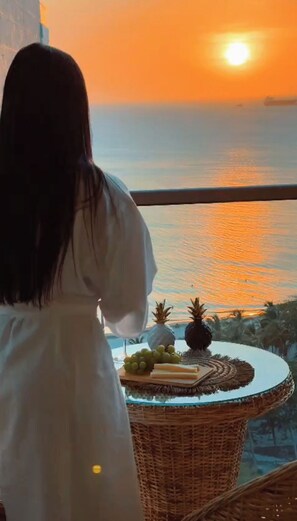 Eine Person in einem weißen Gewand blickt auf einen Sonnenuntergang. Ein Korbtisch mit Früchten und einer Ananas steht daneben.