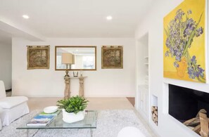 Un salon moderne avec une table basse en verre, un canapé blanc et une cheminée. Deux œuvres d’art encadrées ornent le mur, et une plante en pot est posée sur la table.