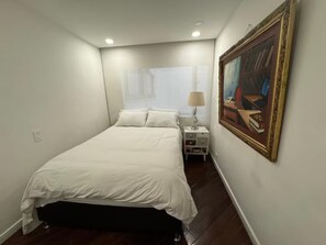 Een slaapkamer met een bed, nachtkastje, lamp en een lijst met een schilderij aan de muur.