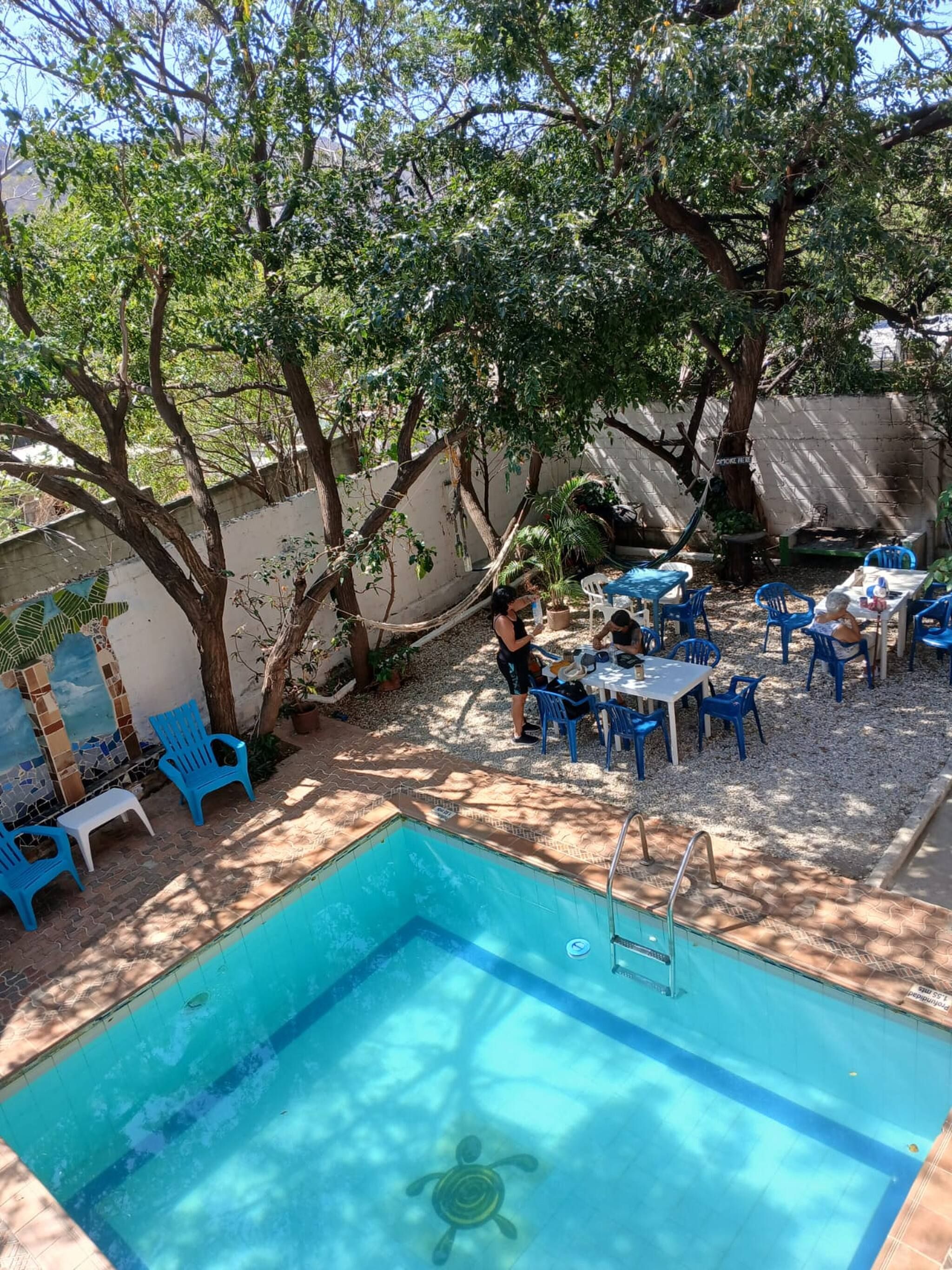 A swimming pool with blue chairs and tables, surrounded by trees and a tiled wall.