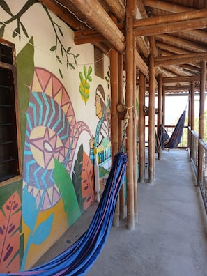 Un espacio al aire libre techado, con pilares de bambú, murales coloridos y hamacas.