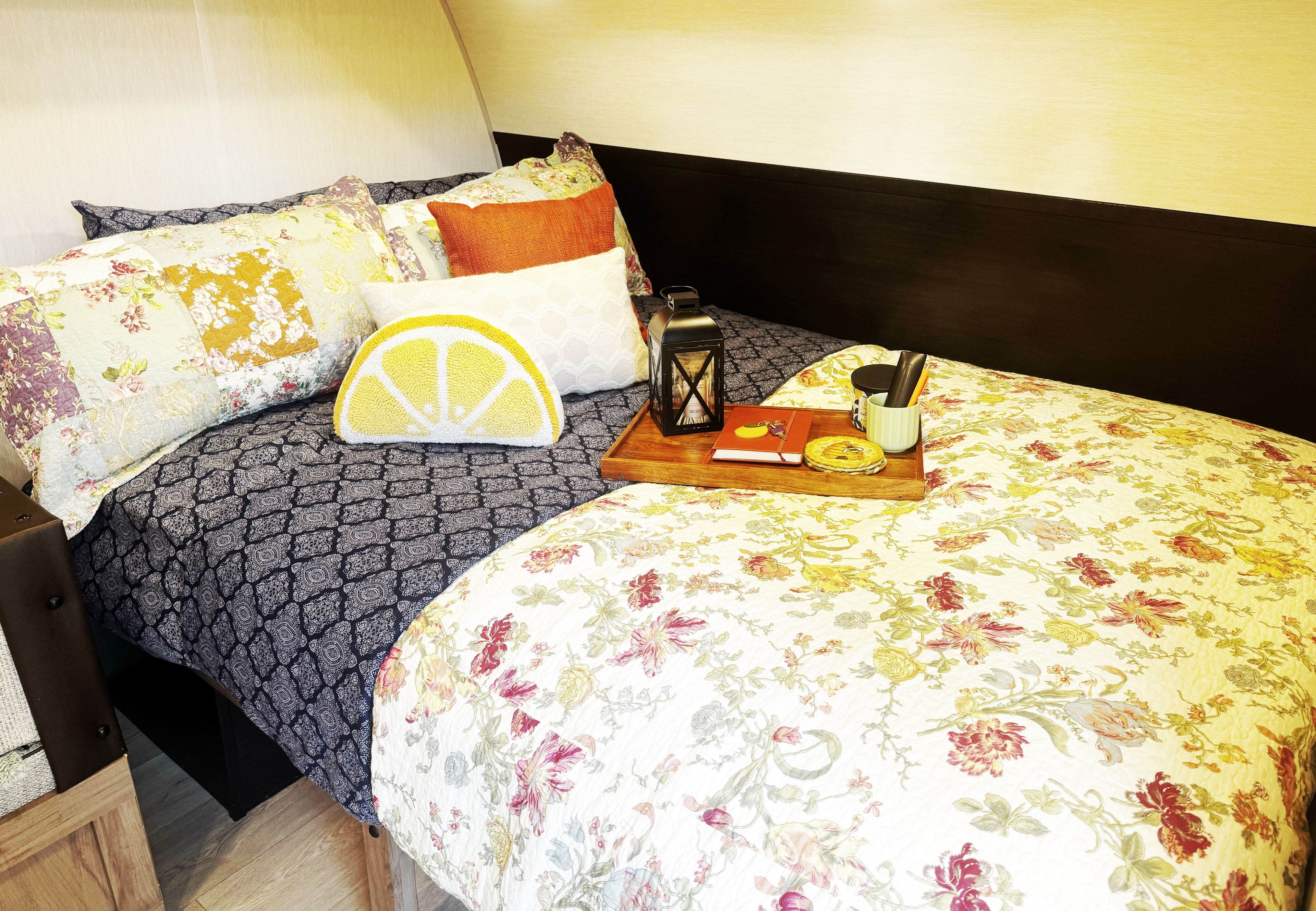 Una cama bien hecha con ropa de cama floral, una mesita de noche de madera, una bandeja con un libro y una vela, y una lámpara de noche.