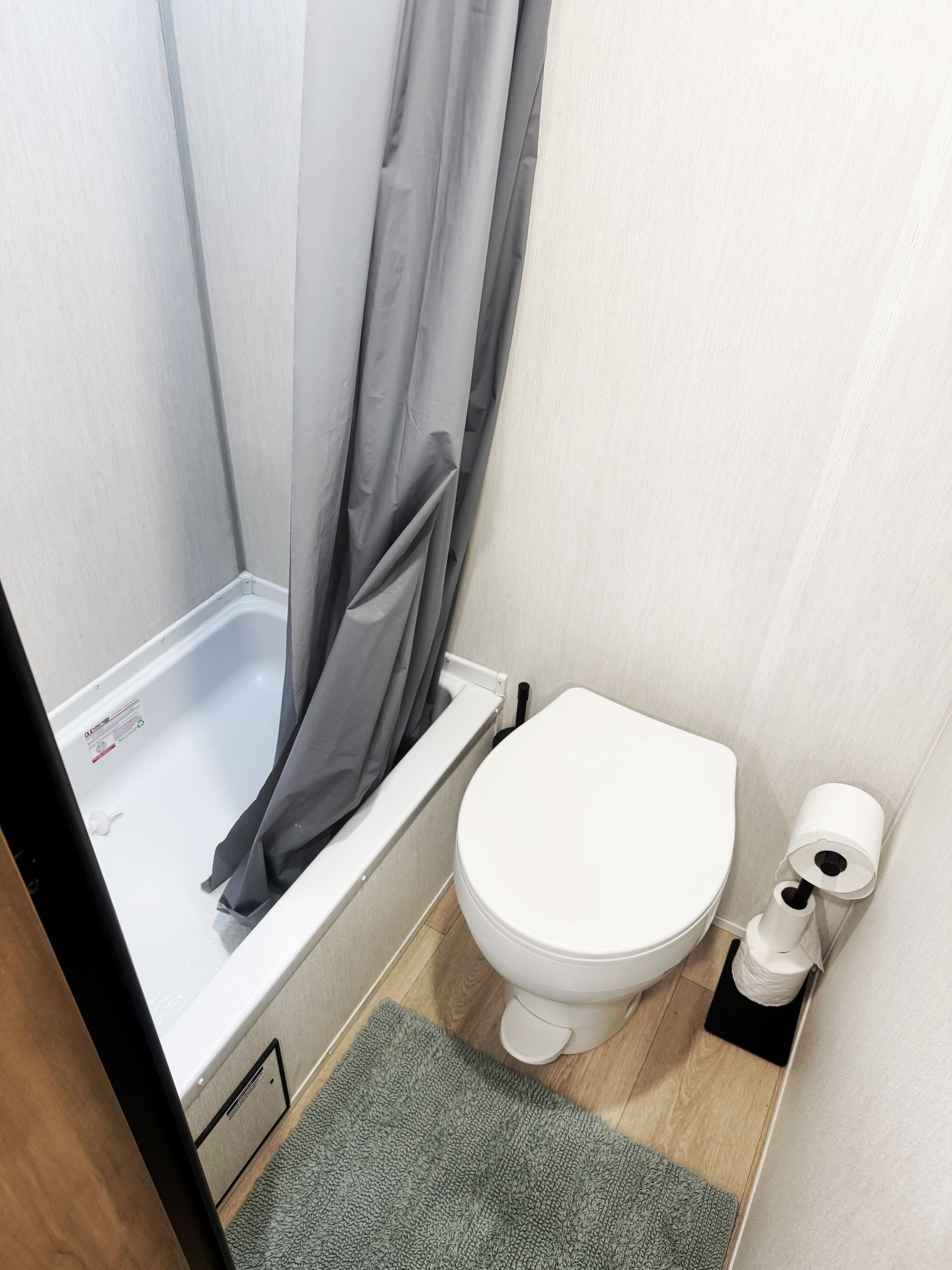 A small bathroom with a white toilet, a shower with a grey curtain, and a green mat on the floor.