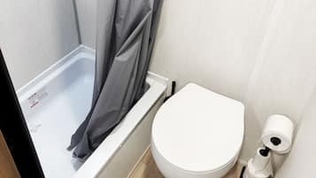 A small bathroom with a white toilet, a shower with a grey curtain, and a green mat on the floor.