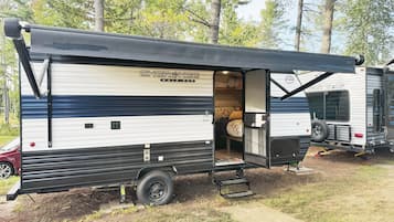 Une maison mobile dotée d’un porche couvert, d’un espace nuit avec un lit et des oreillers, et d’un petit coin cuisine.