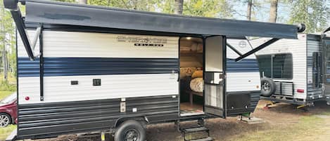 A mobile home with a covered porch, a sleeping area with a bed and pillows, and a small kitchen area.