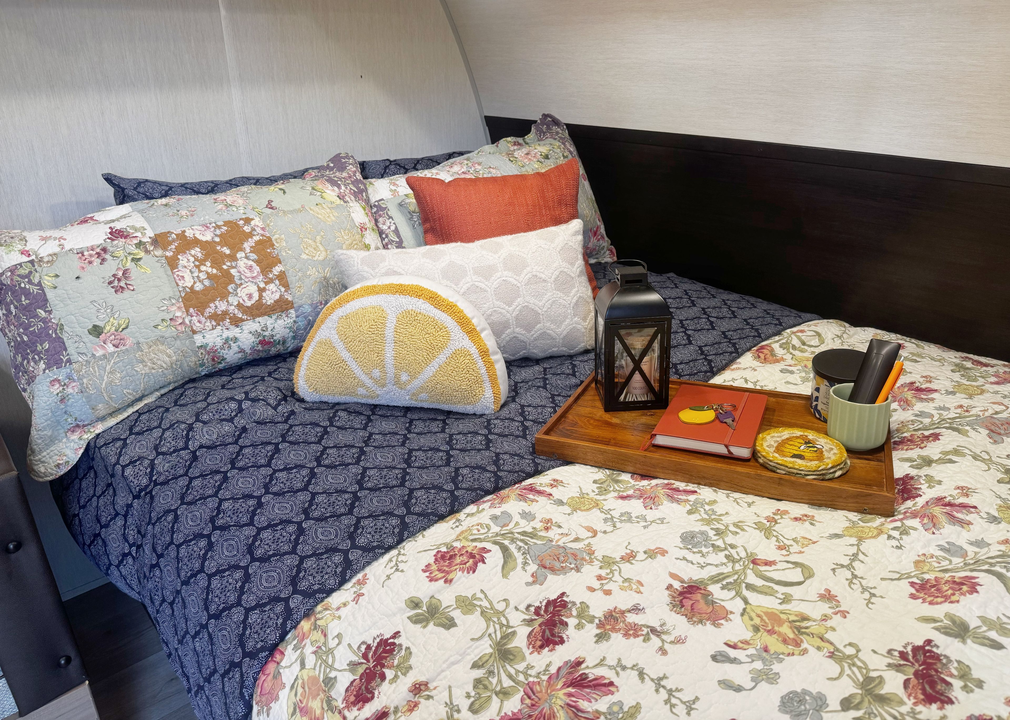 A neatly made bed with floral bedding, a wooden tray with a book, a lantern, and a mug.
