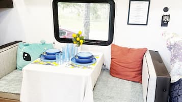 Ein kompaktes Wohnmobil-Interieur mit Esstisch für zwei Personen, einem Fenster mit Blick ins Freie und einer kleinen Sitzecke mit Kissen.
