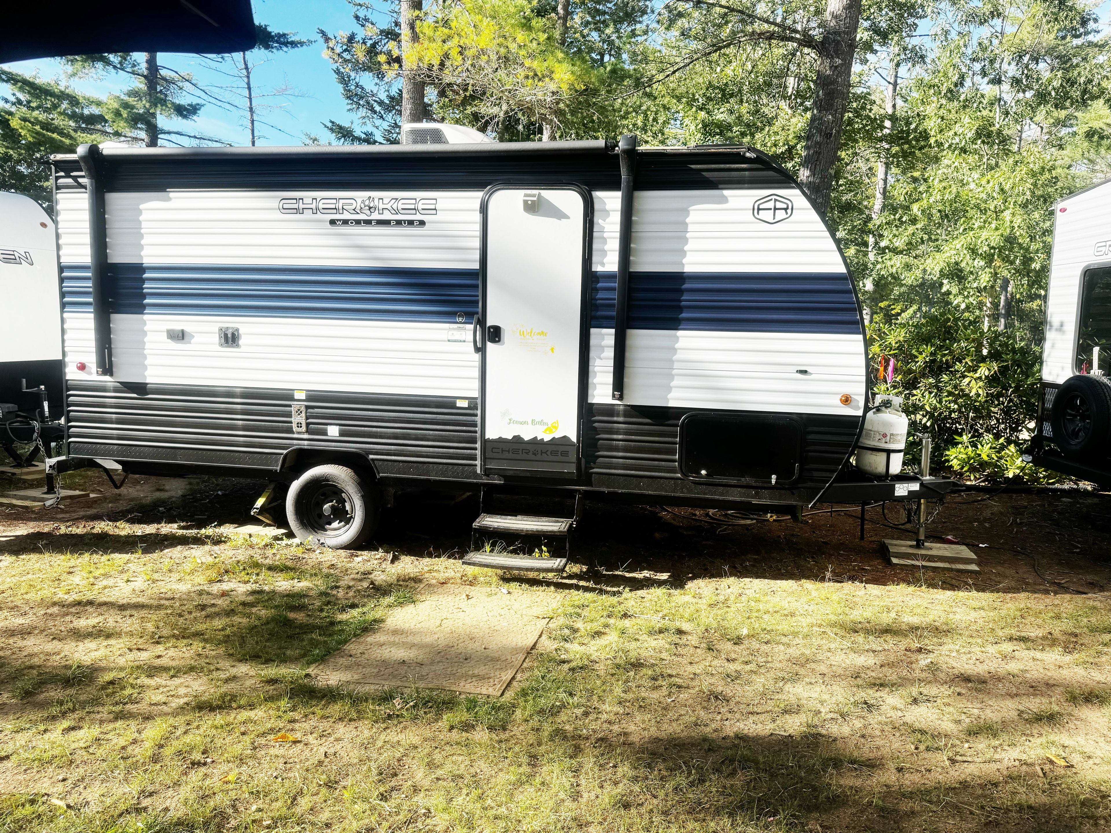 A stationary RV with a visible door and side panel, parked in a grassy area with trees in the background.