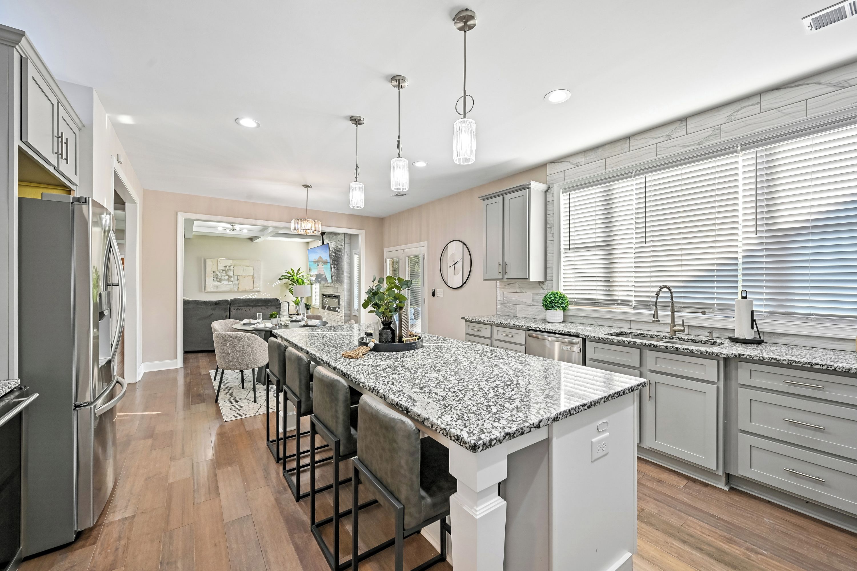 A modern kitchen with a central island, bar stools, pendant lighting, and a large window with blinds.