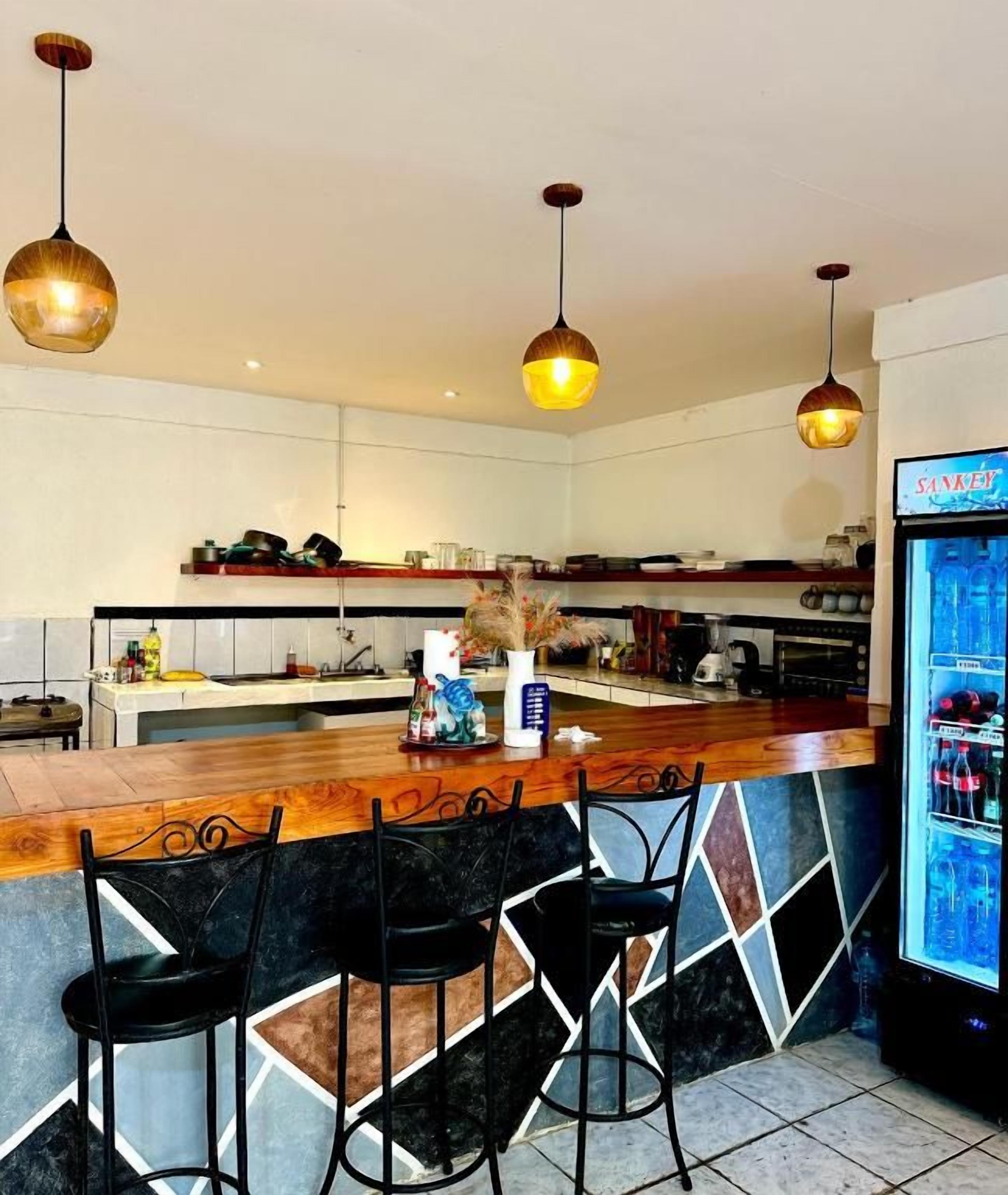 Uma área de bar com balcão e banquetas, geladeira com bebidas e iluminação pendente.