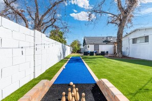 A backyard with a blue lawn bowling area, a white brick wall, and a house with a garage. - Mesa Harmony Haus | Bowling, Beats & Bubbles! (Mesa)