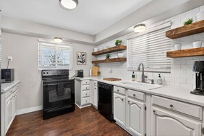 A modern kitchen with white cabinetry, a black oven, a dishwasher, and wooden shelves. - Mesa Harmony Haus | Bowling, Beats & Bubbles! (Mesa)