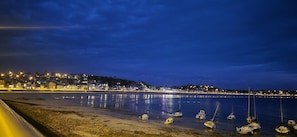 A coastal scene with a calm sea, a sandy beach, and a town with illuminated buildings in the background. - Á Folia - PLENEUF VAL ANDRE (PLENEUF VAL ANDRE)