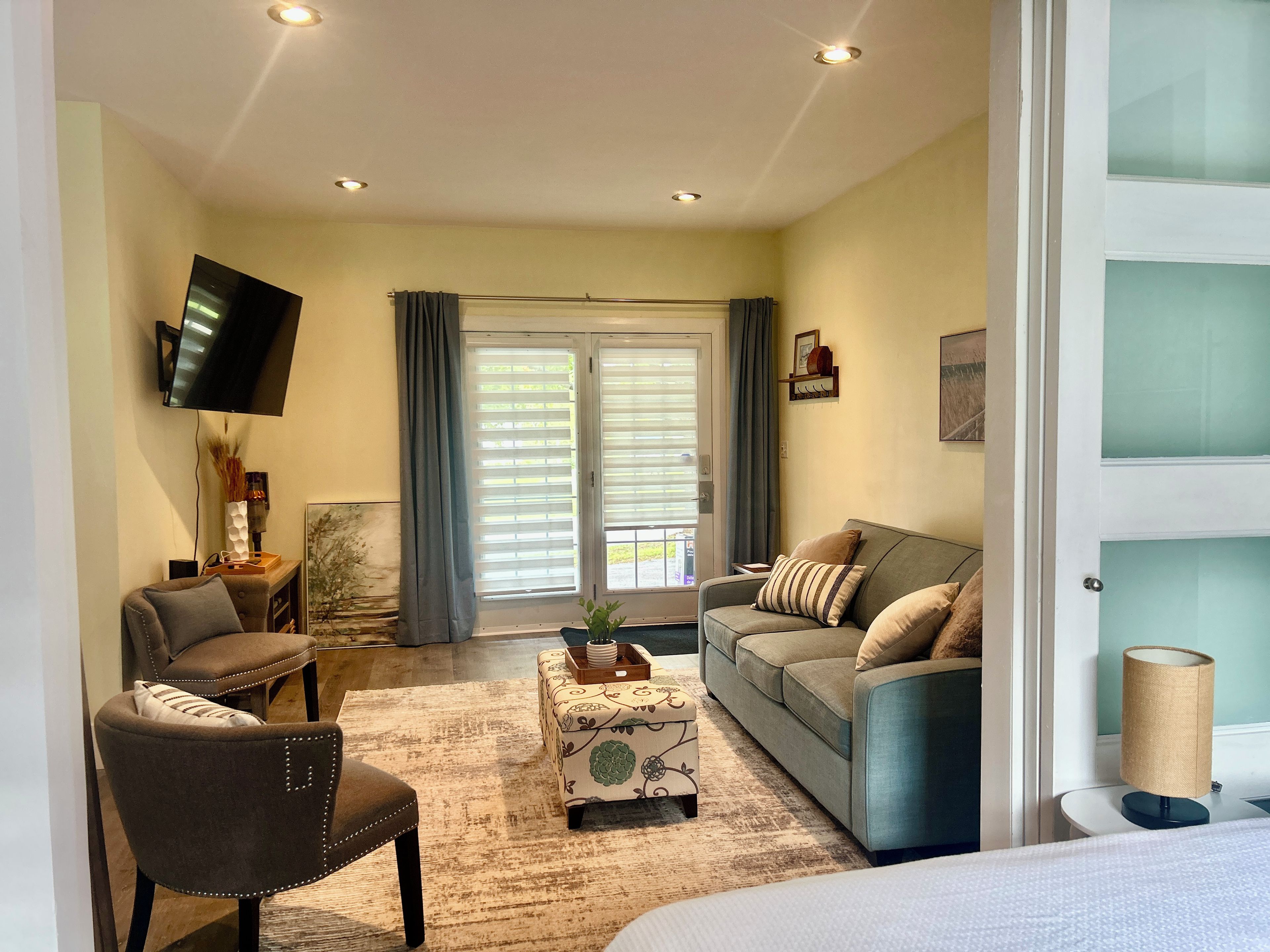 A living room with a sofa, armchair, and ottoman, a flat-screen TV mounted on the wall, and a window with curtains.