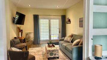 A living room with a sofa, armchair, and ottoman, a flat-screen TV mounted on the wall, and a window with curtains.