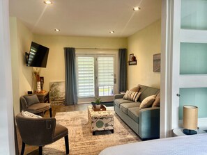 A living room with a sofa, armchair, and ottoman, a flat-screen TV mounted on the wall, and a window with curtains.