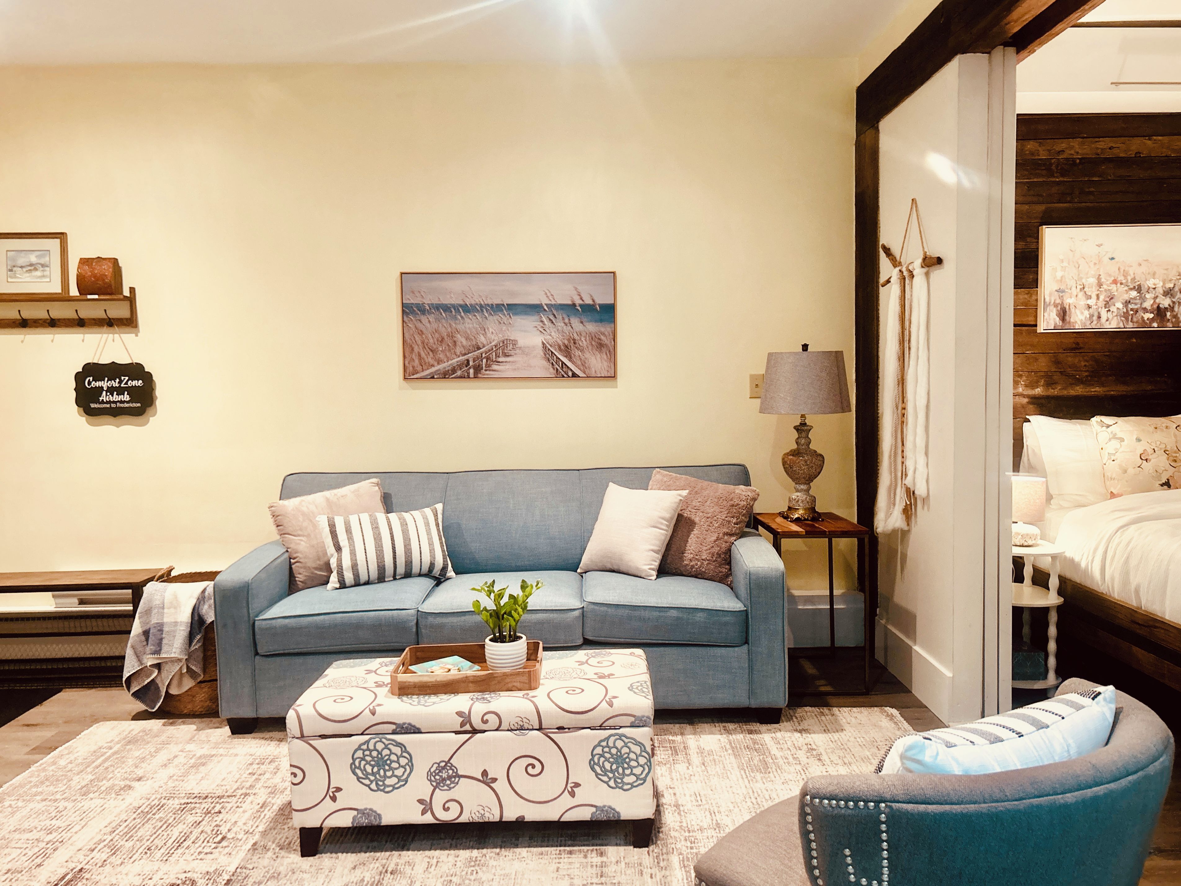 A living room with a sofa, ottoman, and armchair, a lamp, a painting, and a sign on the wall.