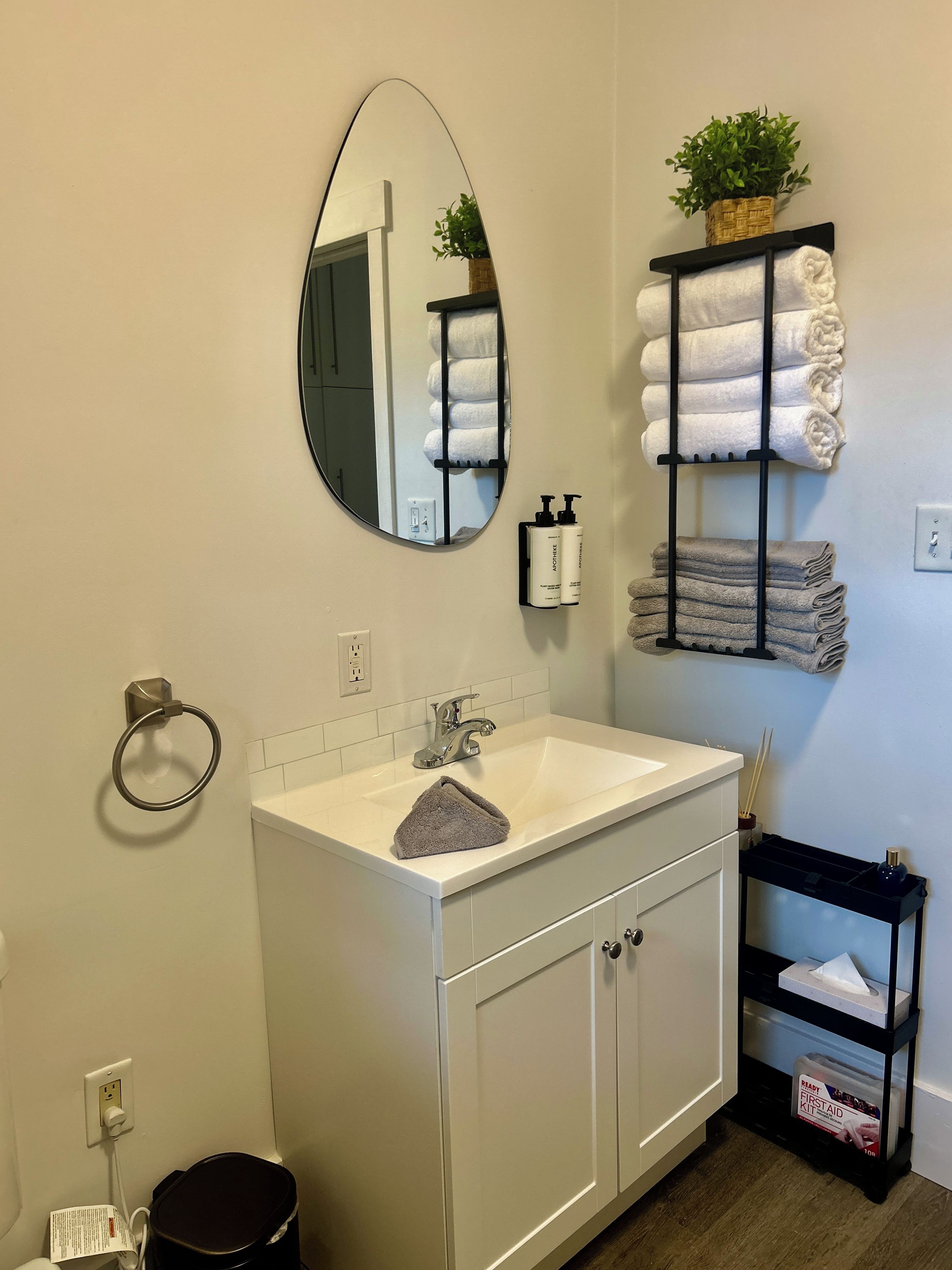A bathroom with a white vanity, a round mirror, a towel rack with folded towels, a small shelf with toiletries, and a black storage unit with a box labeled 'Ready First Aid Kit'.