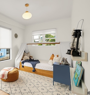 Ein Schlafzimmer mit einem Bett, einer Hängeleuchte, einem Fenster mit Aussicht, einem Nachttisch mit Büchern und einem gemusterten Teppich.